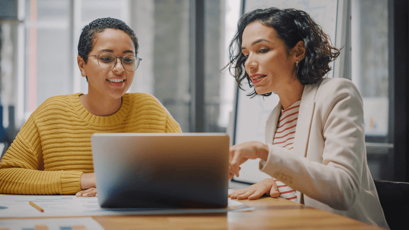 Women at work discussing while looking at a computer screen, main image for blog on the connection between personality assessments and retention rates